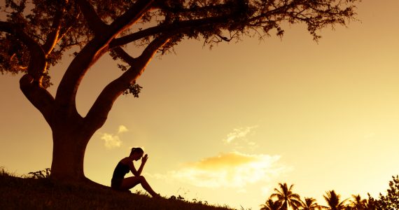 Woman sitting under tree worrying - shadowed image with sunset background