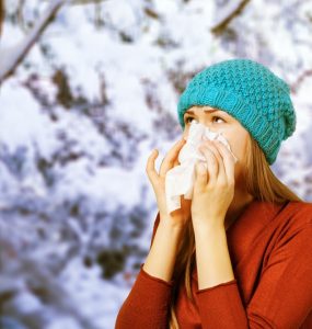 Young woman with the flu wearing a blue woolly hat, blowing her nose with a tissue in a snowy scene
