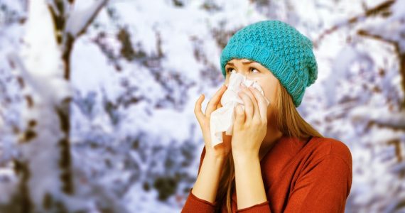 Young woman with the flu wearing a blue woolly hat, blowing her nose with a tissue in a snowy scene