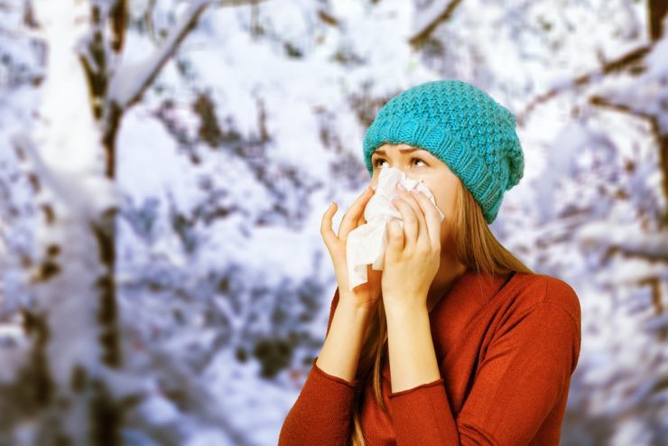 Young woman with the flu wearing a blue woolly hat, blowing her nose with a tissue in a snowy scene