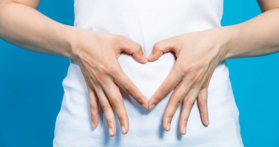 Torso of a woman in white t-shirt making the shape of a heart with her hands, over her stomach.