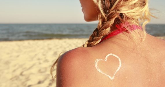 Woman on beach facing the sea with sun scream in a heart shape on her back to represent summer skin