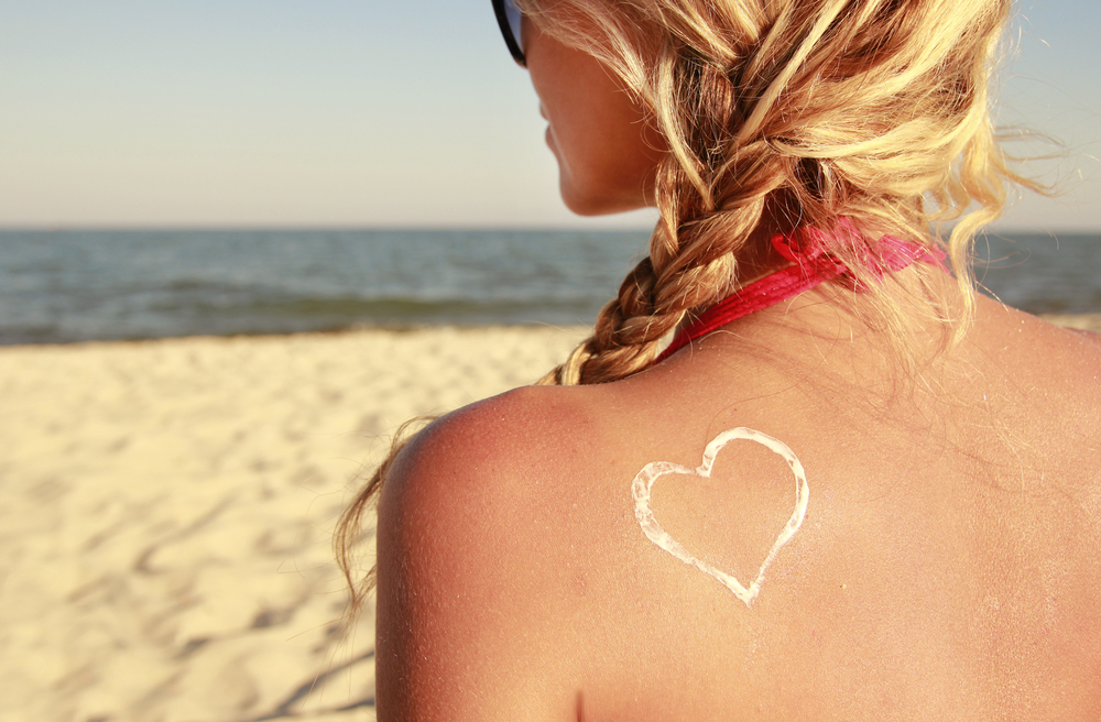 Woman on beach facing the sea with sun scream in a heart shape on her back to represent summer skin