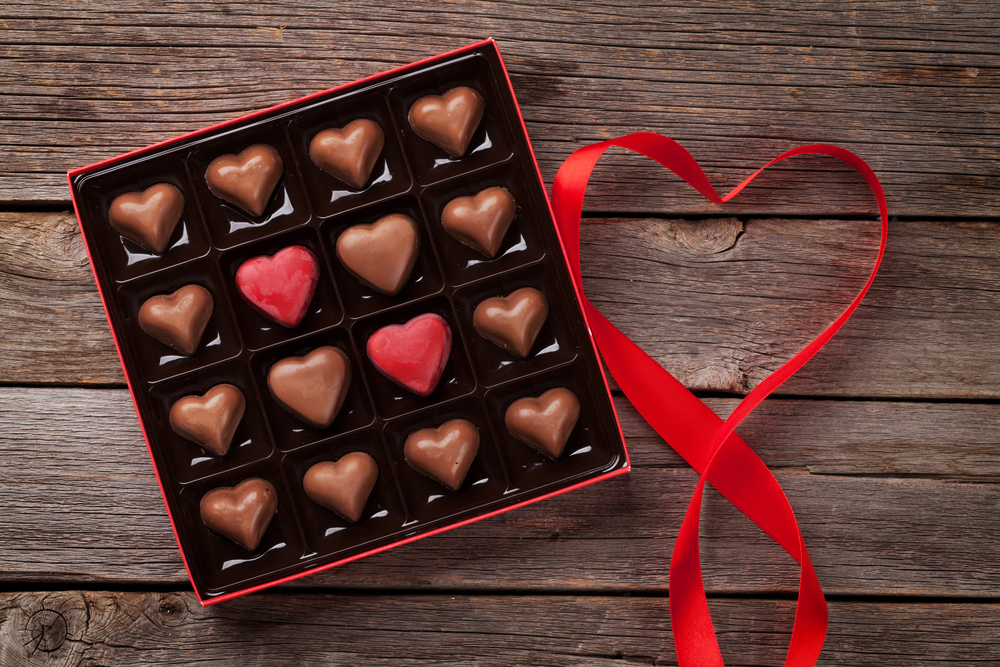 A box of chocolate hearts and a red ribbon in the shape of a heart on a wooden background