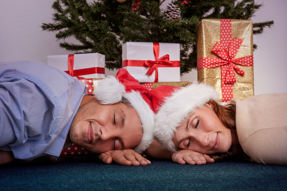 A close up of a man and woman in Christmas hats lyingon the floor with eyes closed to repesent Christmas fatigue and tiredness
