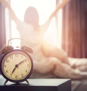 Close up of an alarm clock with a woman rising from bed in the backgruond to represent regular getting up time