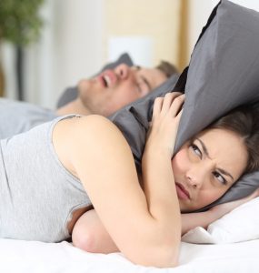 Woman with pillow over her head in bed whilst partner snores