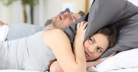 Woman with pillow over her head in bed whilst partner snores